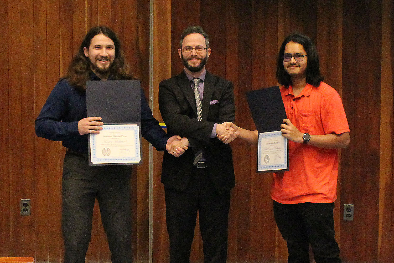Segev BenZvi shakes hands with award winners Hunter Markland and Md Reshad Rahman, each holding their wards.