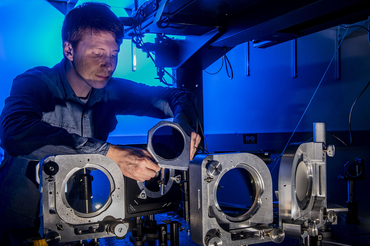 Scientist Jeremy Pigeon installing a radial echelon by inserting a metal part into a system in front of a blue-lit background.
