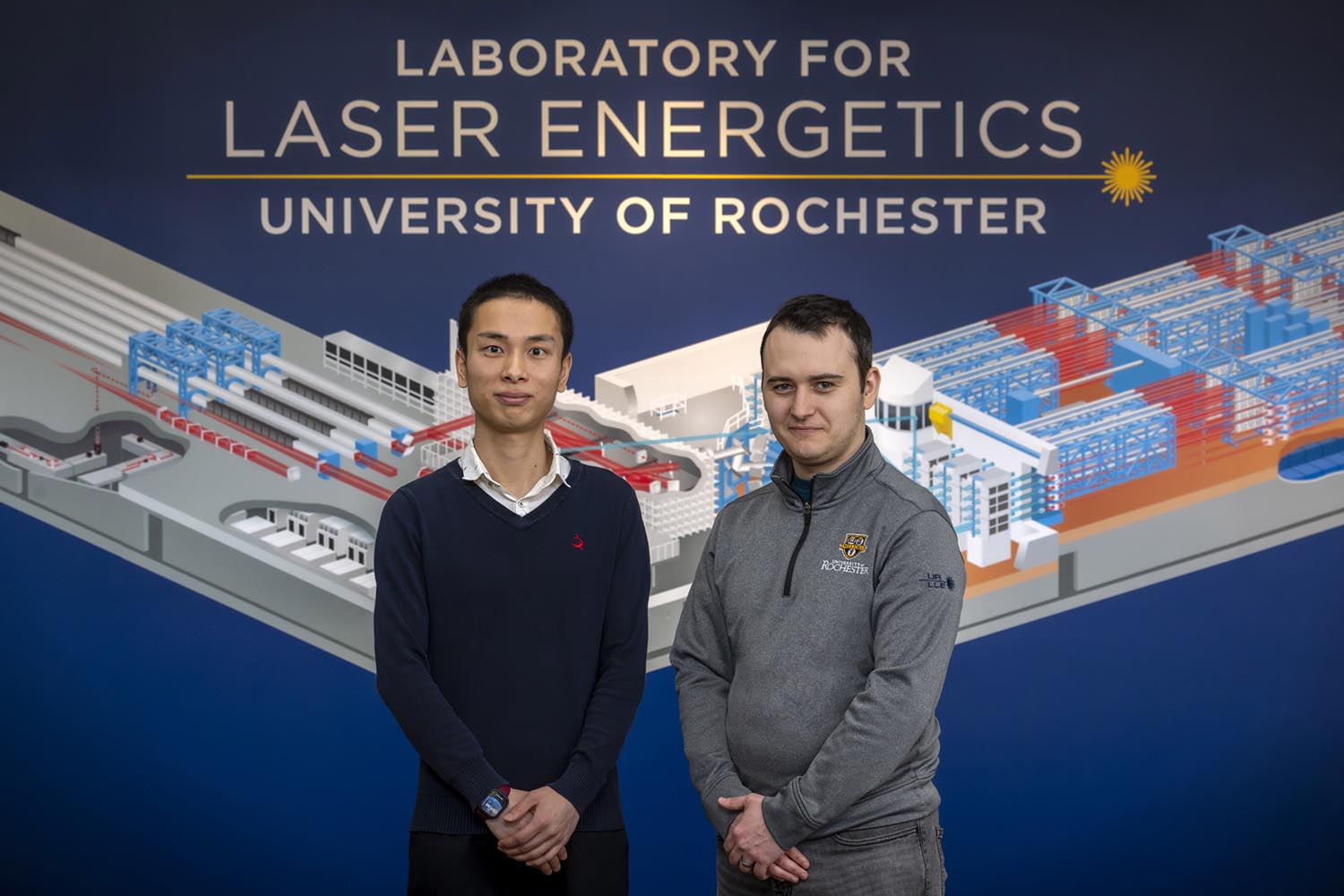 Feature image Victor Zhang and Will Trickey posing in front of the Laboratory for Laser Energetics branding wall.