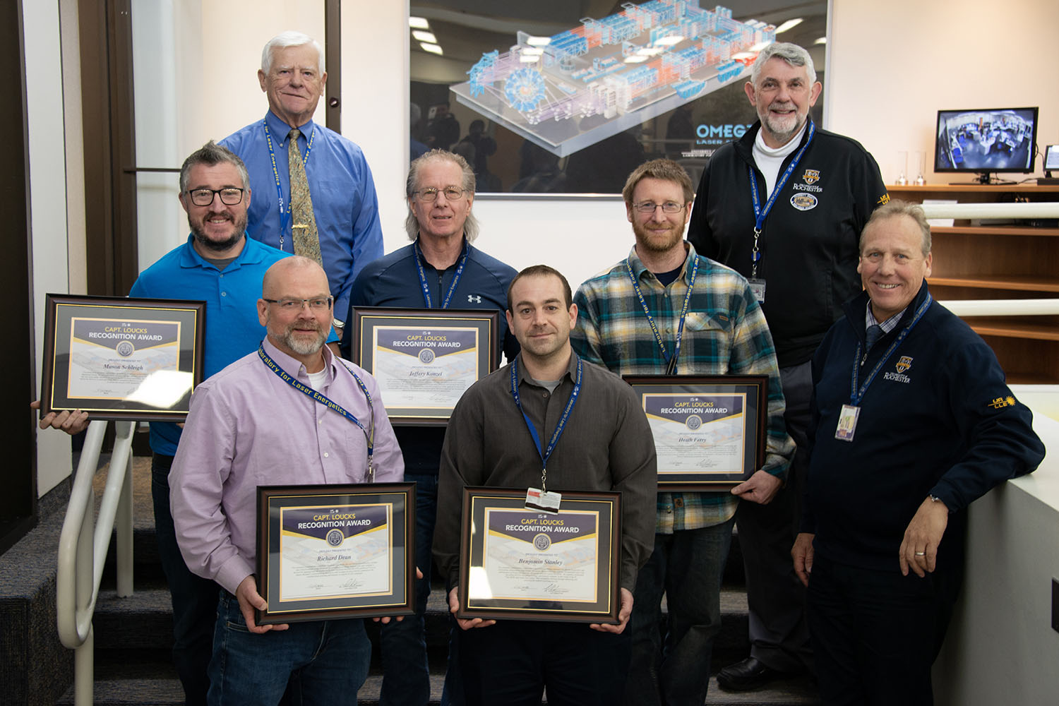 Group photo holding framed awards in front of the OMEGA airbrush illustration.