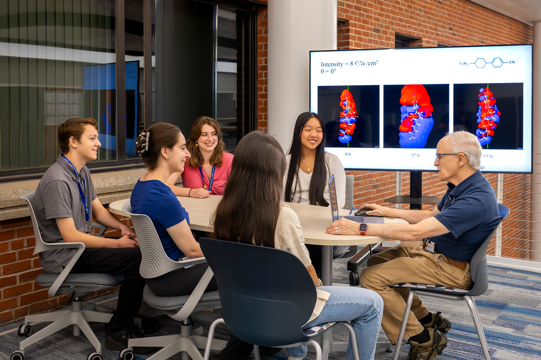 Instructor at a table with students and a monitor showing chemical compounds in the background.