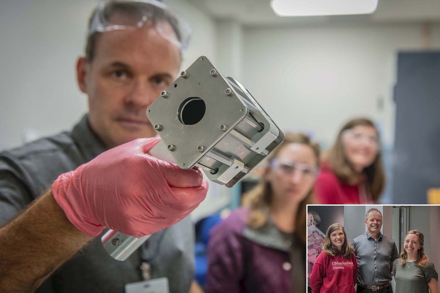 Foreground has a piece of equipment in focus being held up by a researcher. Background are three researchers unfocused in a lab room. Lower right corner in focus are the three researchers.