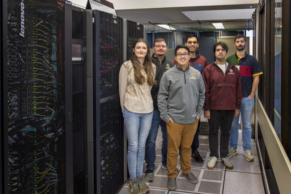 Six people standing next to computer equipment