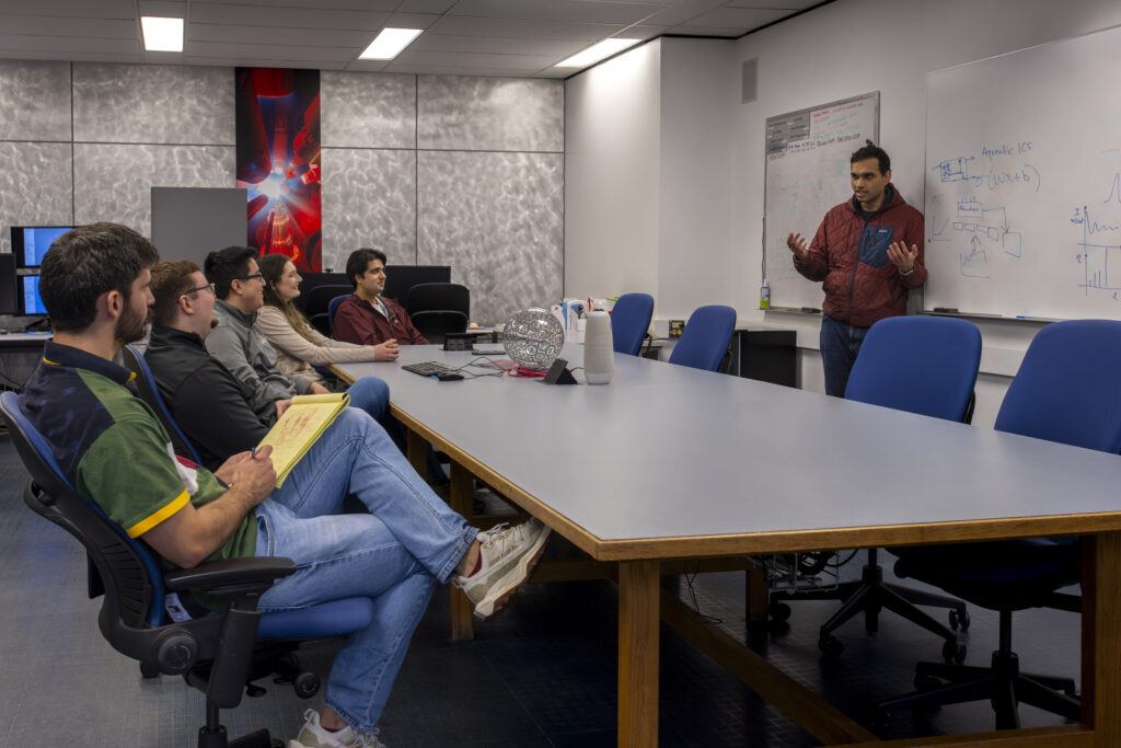 Photo graph of a person standing infront of a white board speaking to people sitting around a conference table.