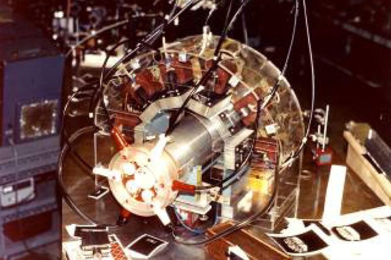 Laboratory setup featuring a cylindrical experimental device surrounded by coils, wiring, and diagnostic sensors mounted on a test stand.