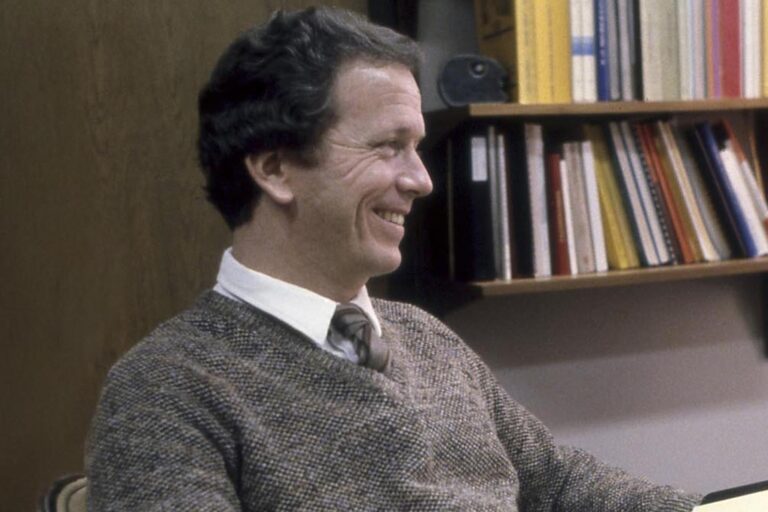 Person smiling while seated in an office with bookshelves in the background.