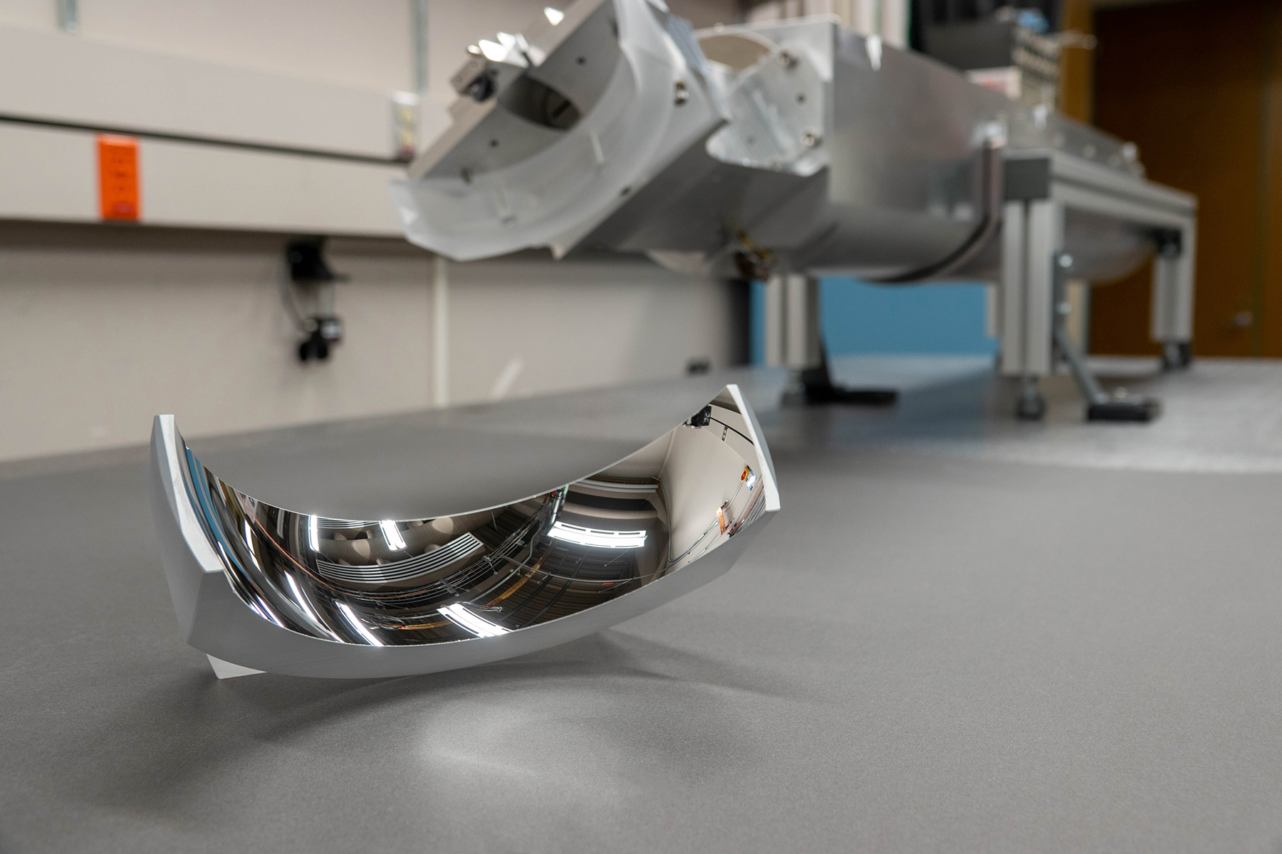 Curved reflective part on a laboratory tabletop with equipment in the background.