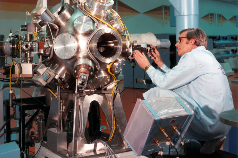 Researcher adjusting instruments on a large stainless-steel vacuum chamber or diagnostic system with multiple ports and cables.