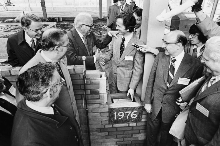 Photo from April 2, 1976 shows several smiling men in dress suits gathered around the building cornerstone which is in engraved with the year 1976. The mood appears celebratory. There are microphones indicative of press coverage.