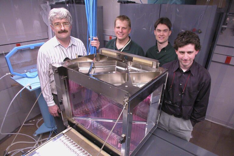 Four people posing by lab equipment in a lab room.