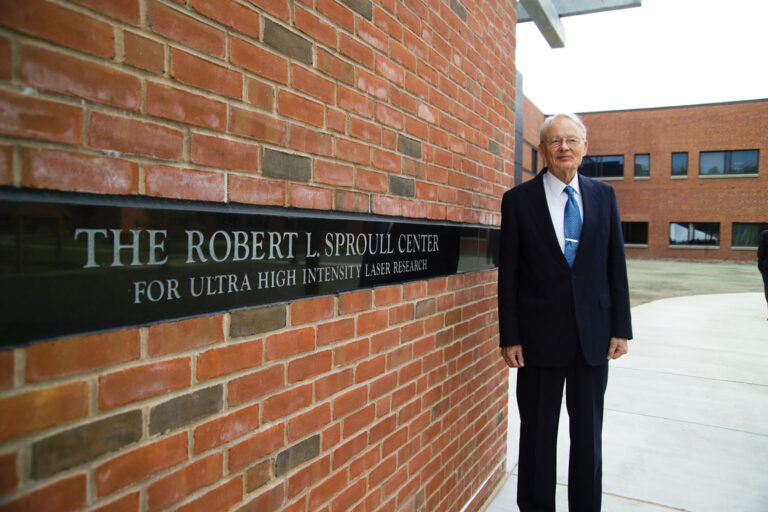 Photo of an older man in dress suit standing in front of a brick wall. The brick wall contains an inscription that reads 
