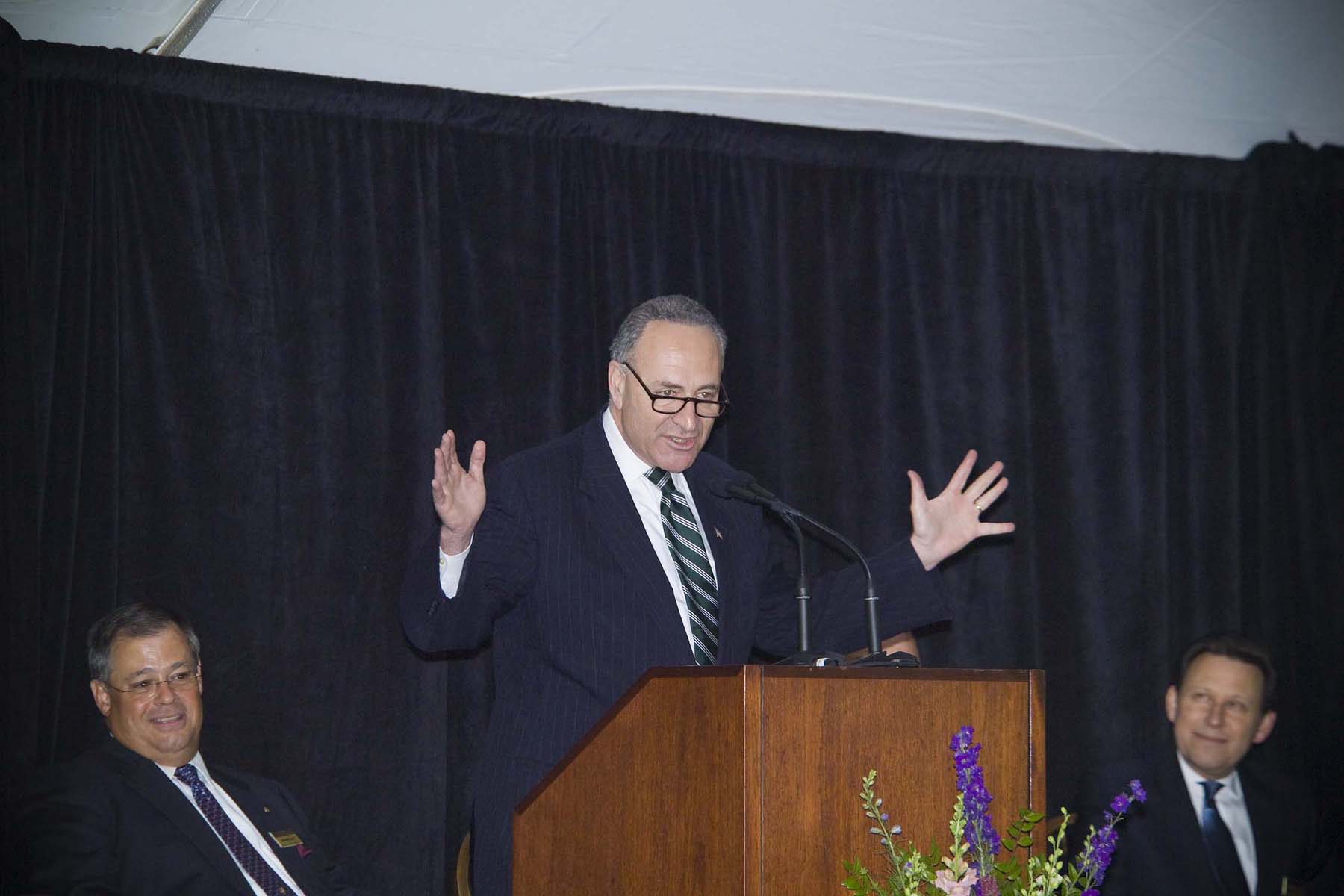 Photo of older man in dress suit gesturing with wide arms at a podium.