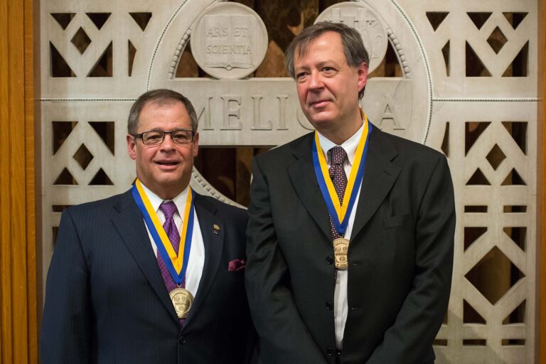 Two people in office attire wearing medals posing in front of wall installation with the word 