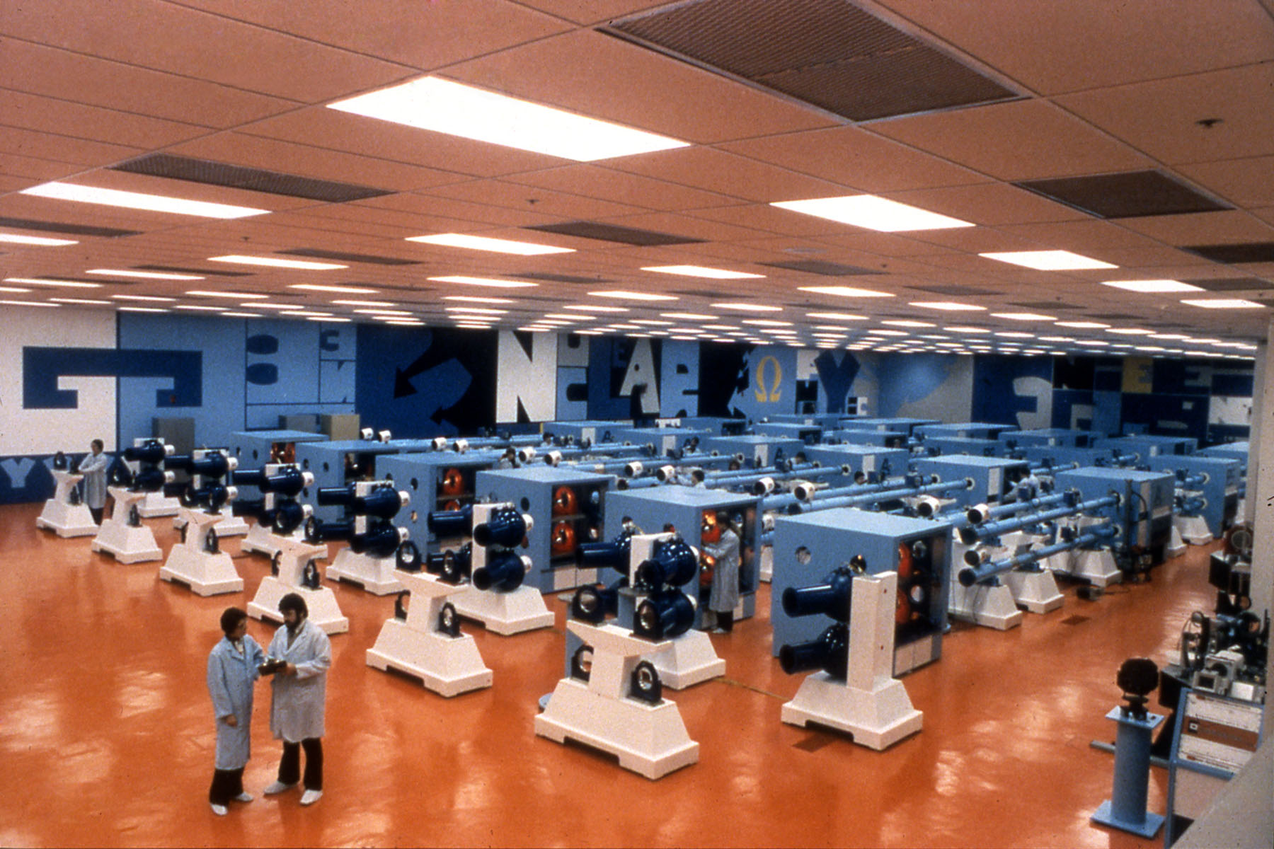 Large laser facility bay with rows of blue laser beamline units and technicians walking through the area.