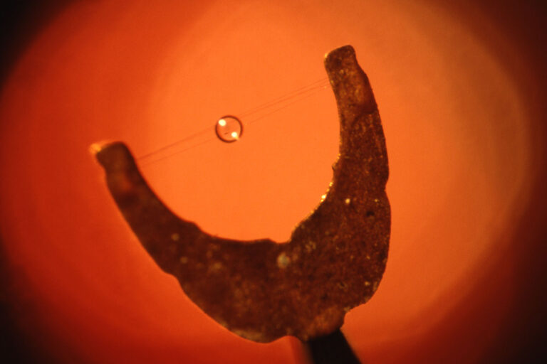 Macro photograph of a small target suspended above a crescent-shaped metal structure, illuminated with a warm orange background.