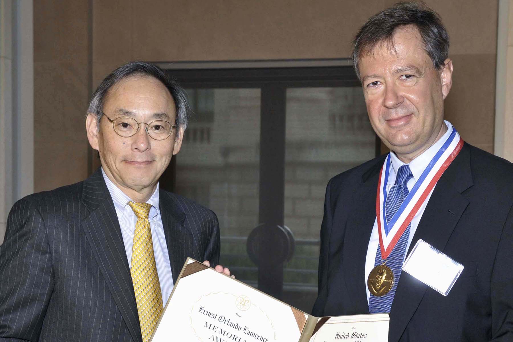Photograph of two men in suit and ties holding framed certificate or award. One of the men weart a medal on a ribbon around his neck.