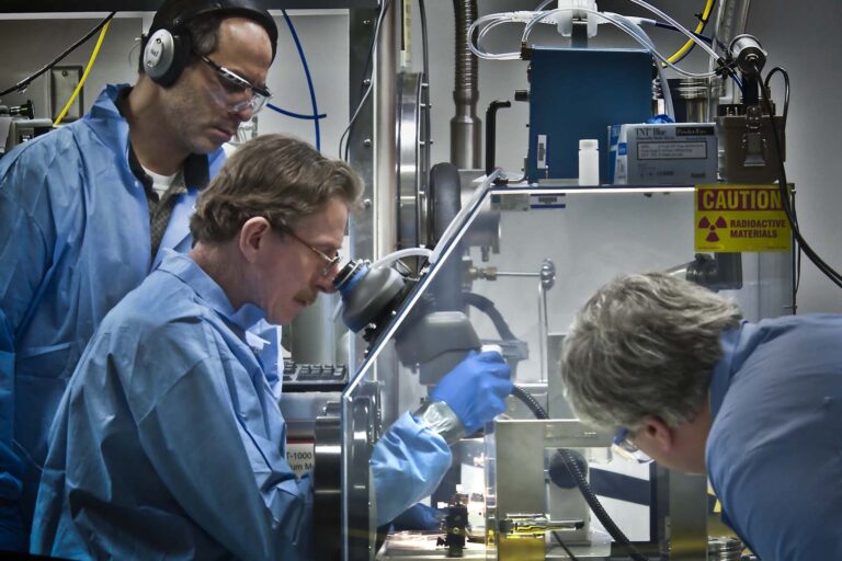 Photo of three scientists or technicians in lab apparel mount inga target. One looks through a microscope and manipulates equipment through a glove box.