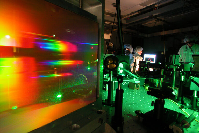 A photo inside a dark lab room with green and rainbow lighting. Lab equipment in the foreground. Lab workers in lab attire shown in background.