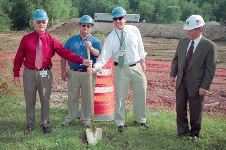 Four men wearing hard hats and holding a golden shovel at a groundbreaking ceremony on a construction site, standing beside an orange safety barrel.