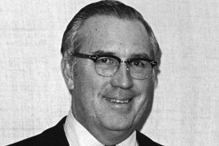 Black-and-white portrait of a man wearing glasses and a suit, smiling slightly.