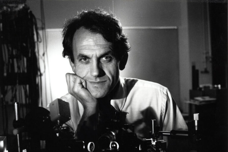 Portrait of a man leaning on scientific equipment, looking into the camera with his hand resting on his face.
