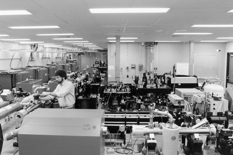 Wide view of a laser laboratory with multiple optical tables, instruments, and researchers working at various stations.