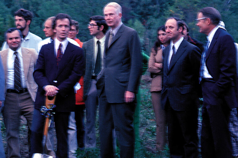 Outdoor photo of several men in suits standing together during a formal event or site visit. One is holding a shovel.