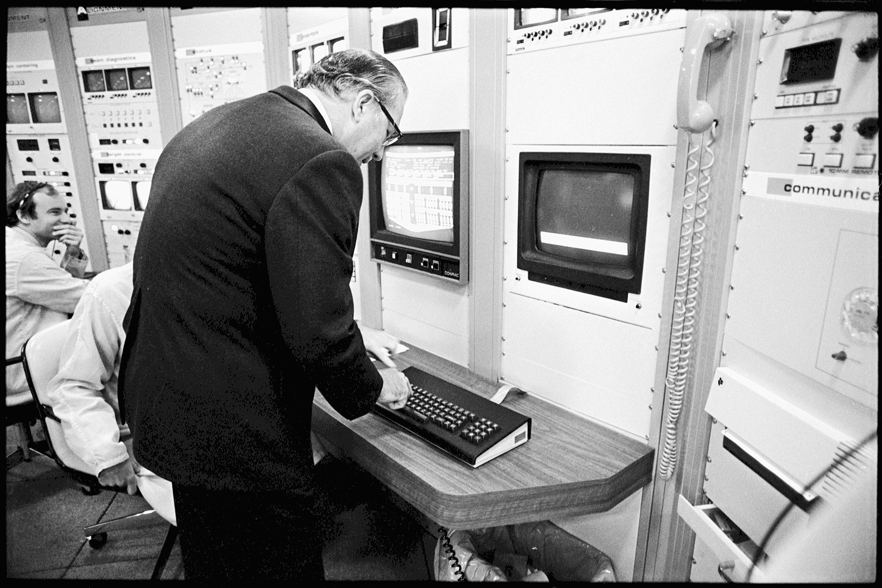 Man operating early computer or instrumentation equipment, standing at a console with monitors and control panels.