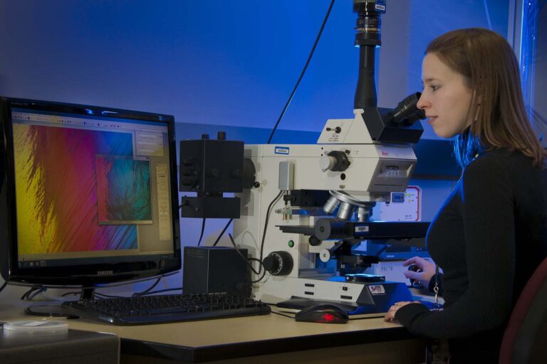 Photo of a scientist in a lab looking through a microscope. A computer monitor sits next to the microscope.