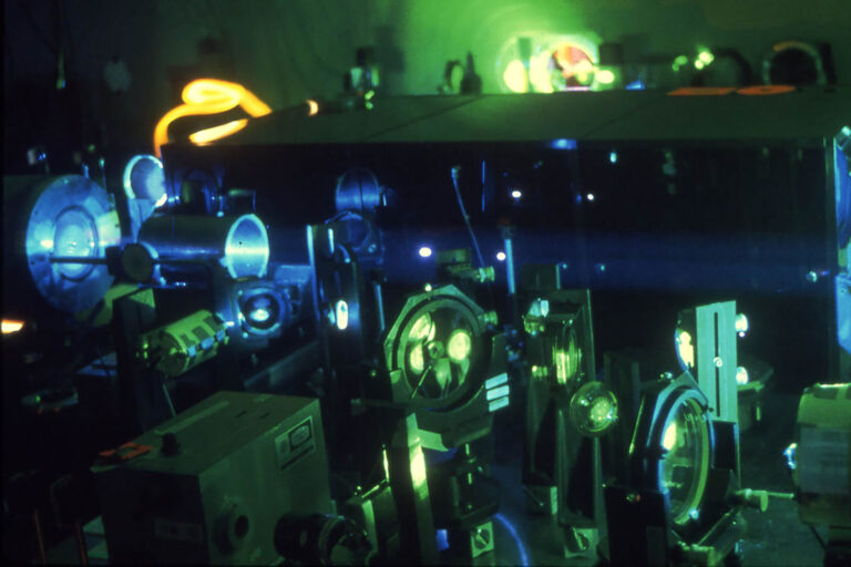 Laser laboratory scene with optical components and beamlines illuminated by bright green and yellow light.