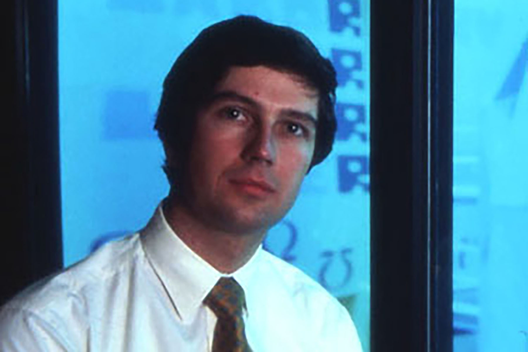 Portrait of a man in a white shirt and tie standing in front of a blue background with scientific graphics.