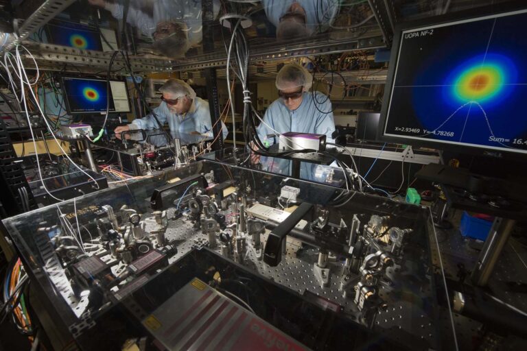 Two scientists in cleanroom apparel and protective eyewear make adjustments to an amplifier.