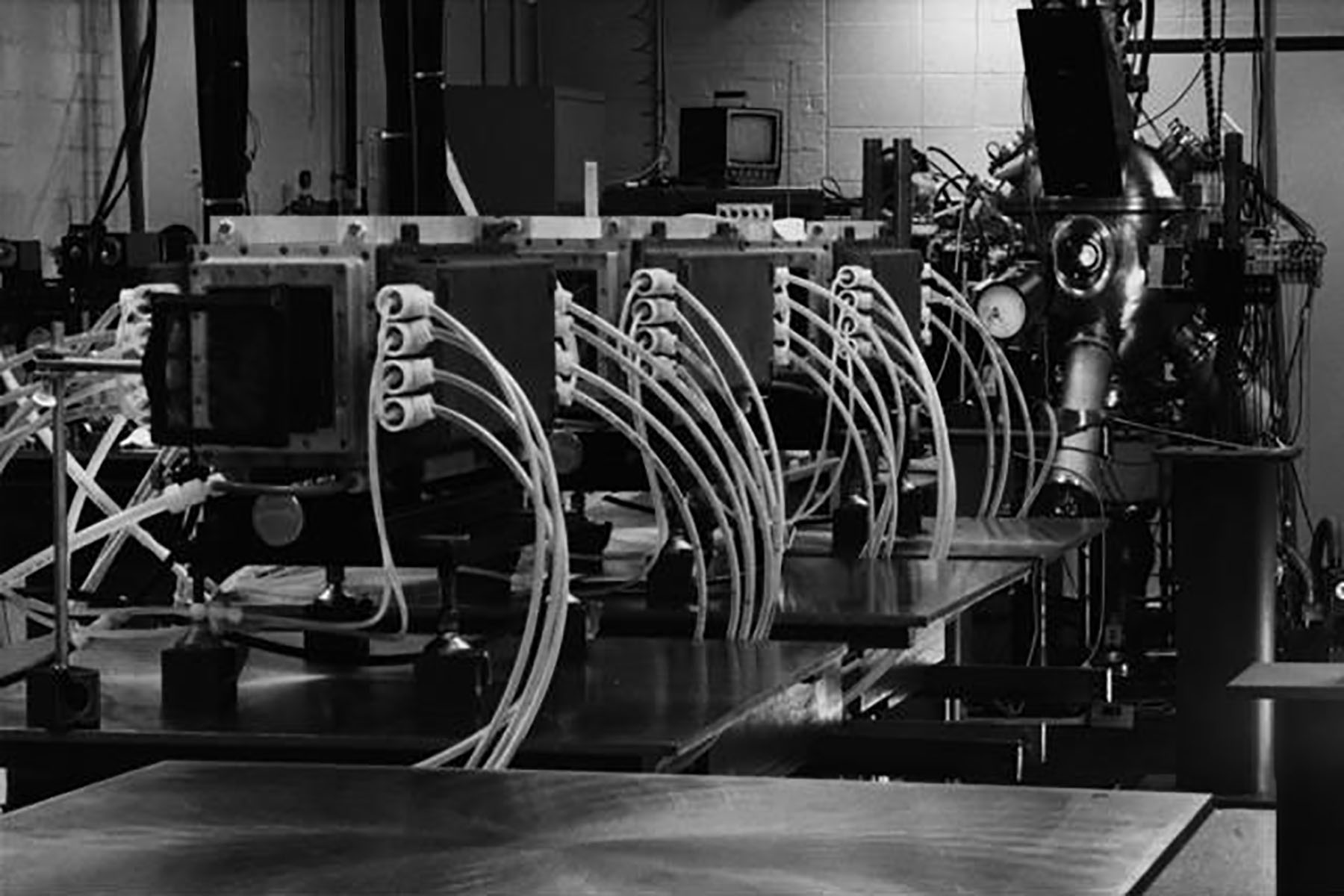 Early laser laboratory equipment featuring a row of large optical components connected by arched cables or tubing.