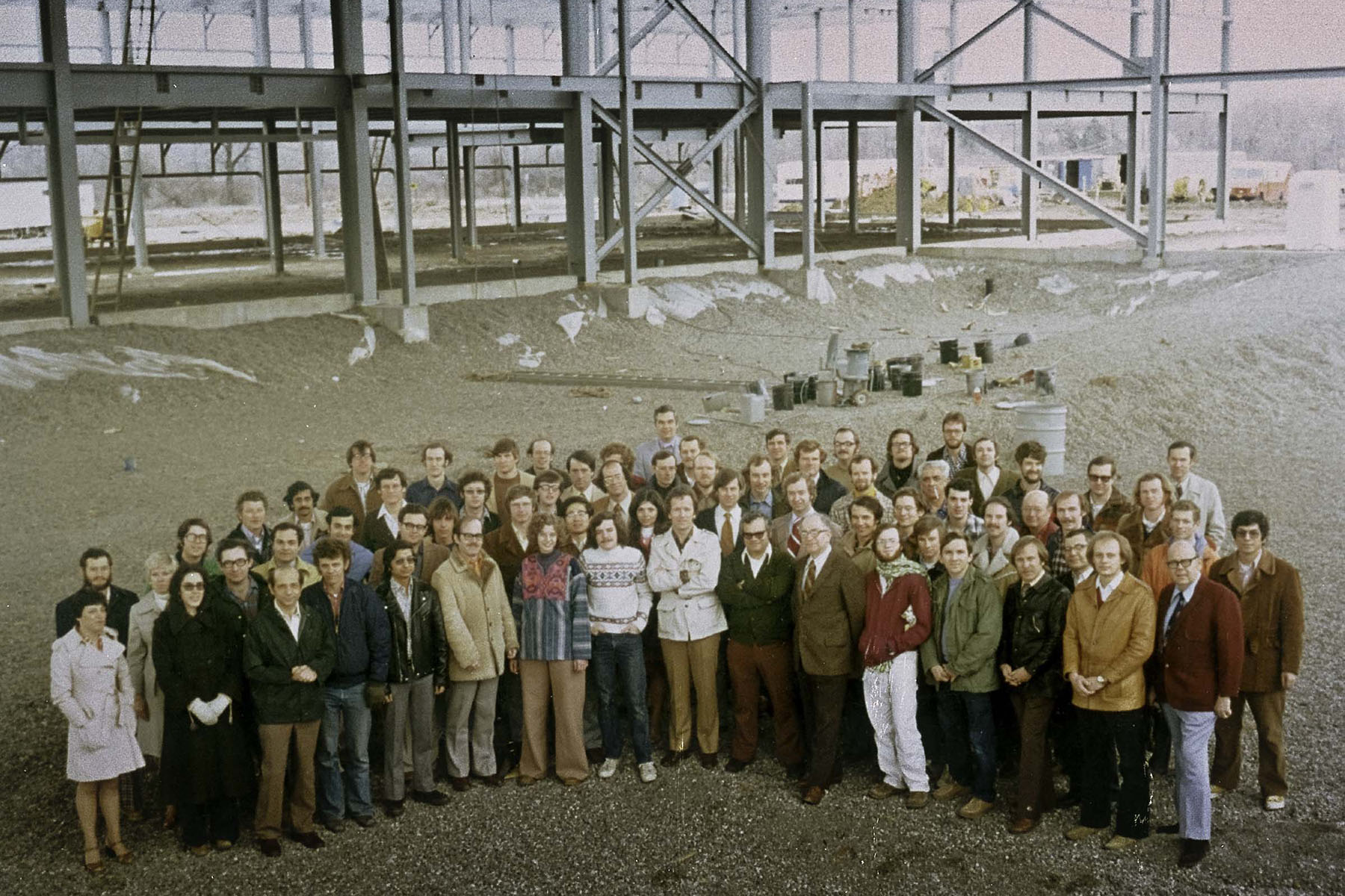 A group of 60-70 people stands in front of the bare framework of the LLE building in Rochester, NY. The surrounding area is bare and there are light snowdrifts on the ground.