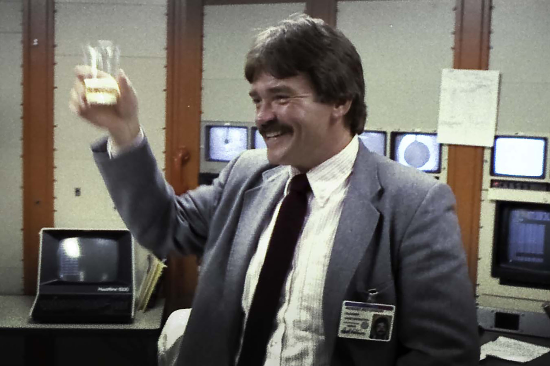 A man in a suit smiling and holding up a glass as if to toast while standing in a laboratory environment.