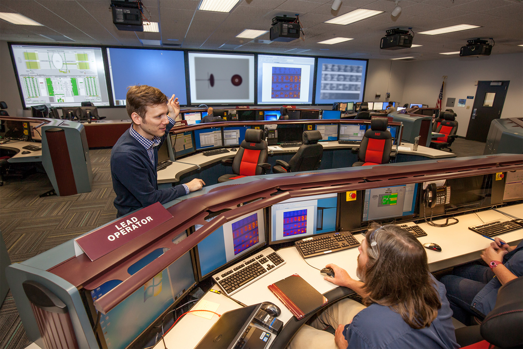 Students and researchers work at computer stations inside a modern control room with multiple monitors and large display screens on the walls.