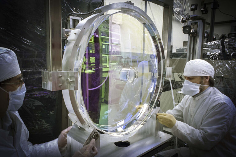 Technician in cleanroom attire adjusts a large focusing parabola lens for the LLNL Petawatt Laser during final photometric testing in LLE’s OMAN Shop.