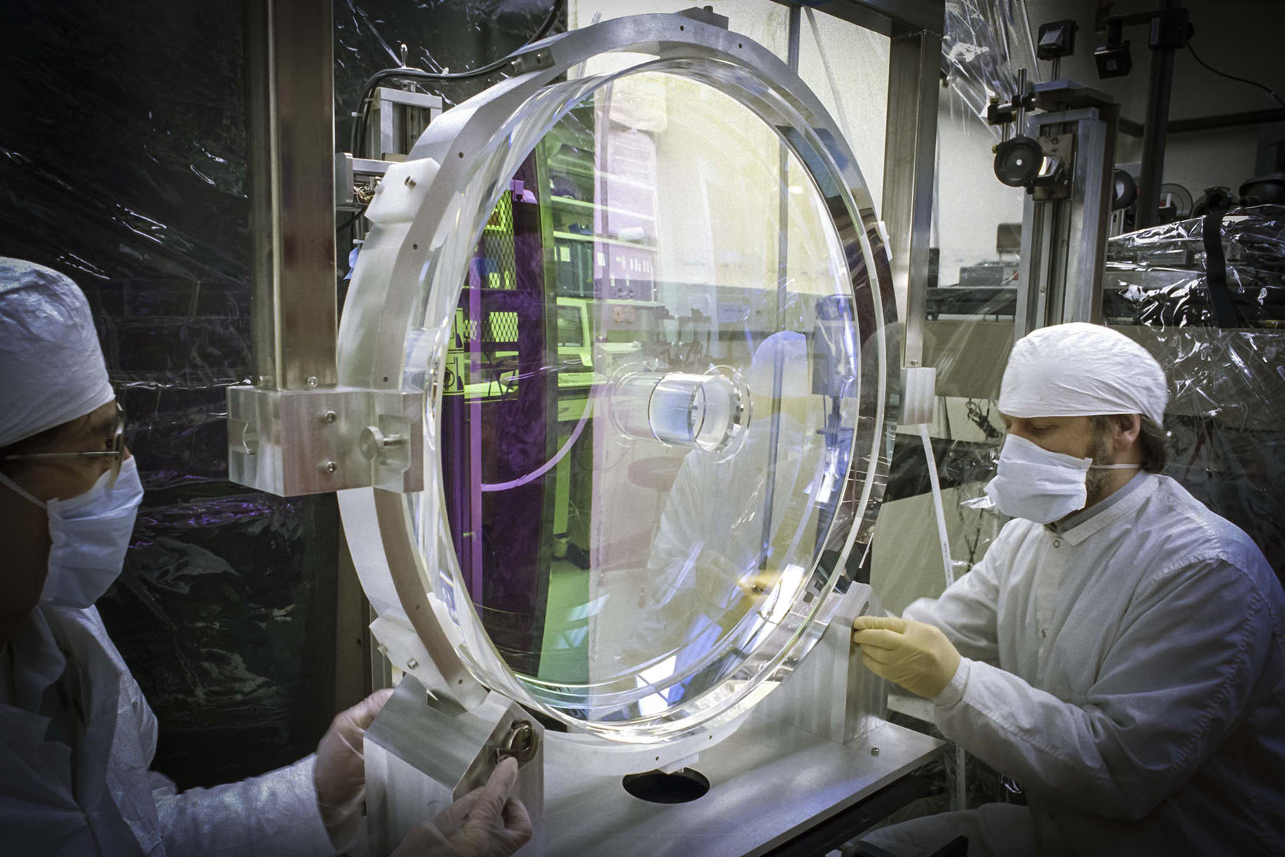 Technician in cleanroom attire adjusts a large focusing parabola lens for the LLNL Petawatt Laser during final photometric testing in LLE’s OMAN Shop.