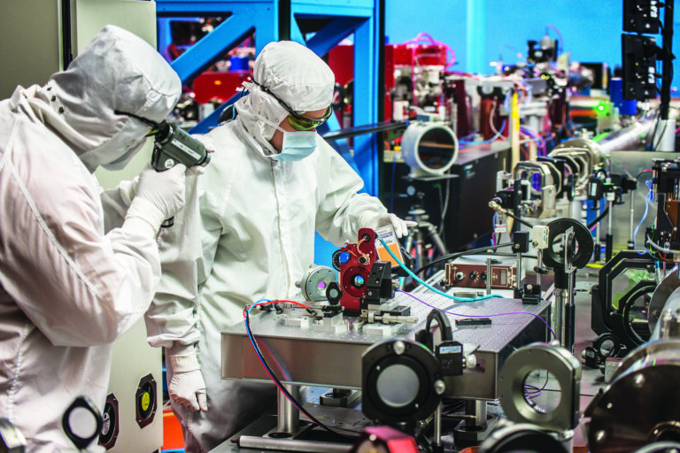 Technicians wearing white cleanroom suits working with complex optical and laser equipment in a brightly lit laboratory.