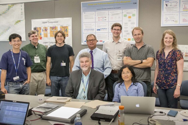 A group of nine people in a meeting room posing for a photo, sitting and standing around a conference table with laptops, notebooks, and posters behind them.