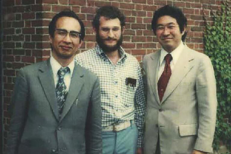 Three people standing together outdoors in front of a brick wall, all dressed in collared shirts and jackets.
