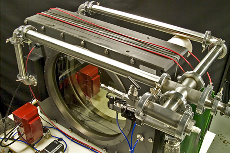 A close-up of a high-precision optical instrument with metal tubing, alignment hardware, and a large circular glass window—part of a laser diagnostic or vacuum system.