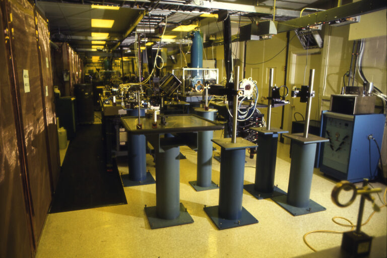 Laser laboratory setup with multiple optical tables and pedestal-mounted optics arranged in sequence.