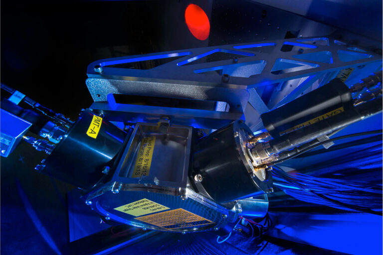 Close-up of a blue-lit laser optics system showing precision mirrors, beam alignment modules, and safety warning labels.
