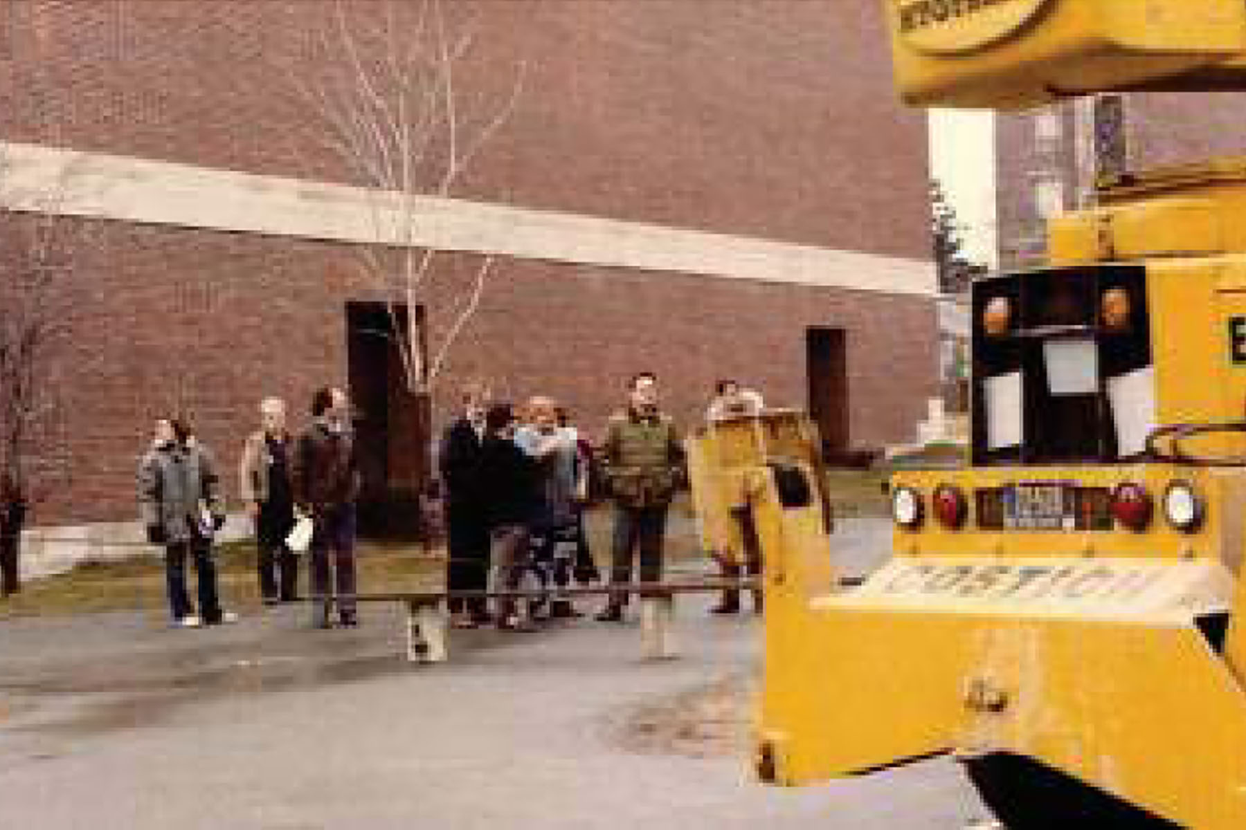 A group of people gathered outside a brick building observing or assisting with construction work near a large piece of yellow machinery.