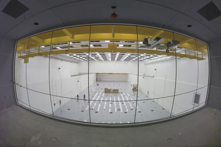 Photo from inside the viewing gallery with windows overlooking a lab room containing several construction cones, equipment, material, and two workers.