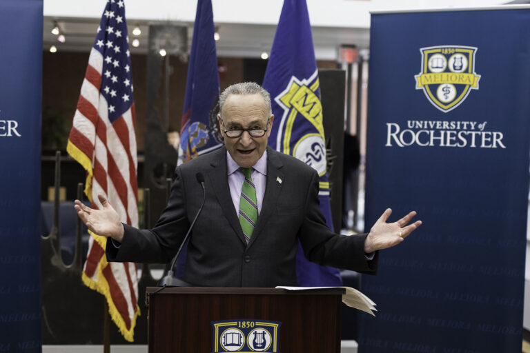 A man speaking at a podium with flags of the US, NY state, University of Rochester and a University of Rochester banner behind him.