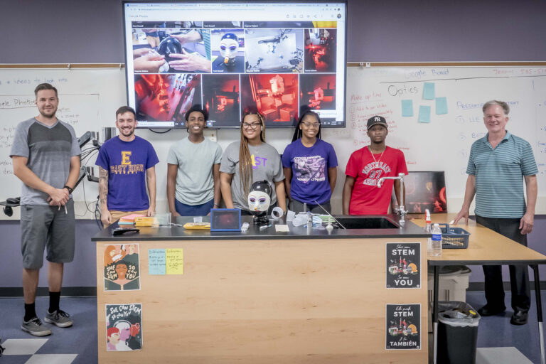 Group of students standing behind a demonstration table with robotics and science equipment, posing for a photo in a classroom.