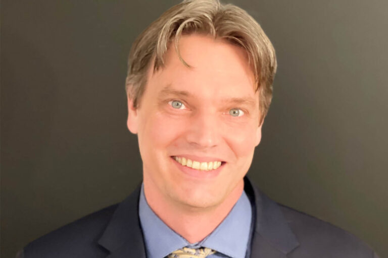 Portrait of a smiling man wearing a suit jacket, dress shirt, and tie, photographed against a neutral background.
