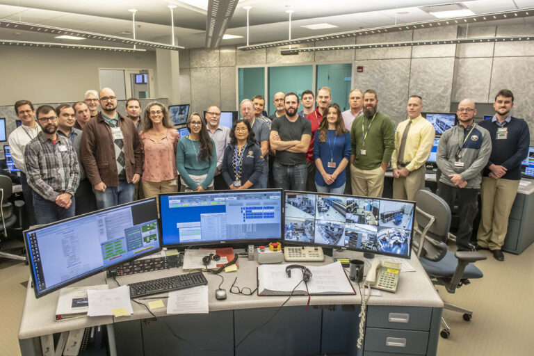 Group of people standing in a control room behind several computer monitors displaying live experimental feeds.